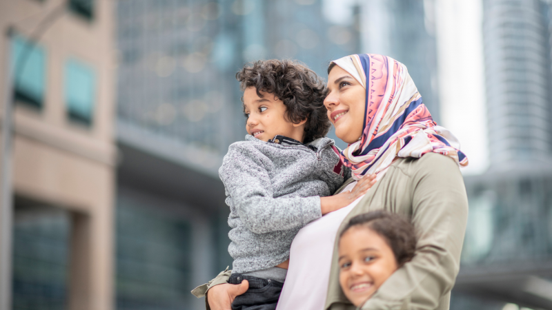 Image features a mother with two children, standing on a city street, one child stands next to her and one is in her arms. They are all looking away and smiling.